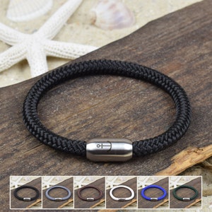 Könnte beinhalten: Ein schwarzes Armband aus geflochtenem Seil mit einem zylindrischen Magnetverschluss aus Edelstahl, der mit einem kleinen Ankersymbol graviert ist. Das Armband wird neben einer Miniaturansicht gezeigt, die weitere Farboptionen wie Grau, Braun, Weiß, Blau und Grün darstellt.