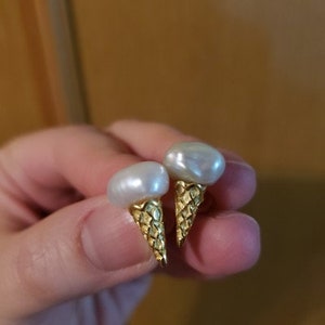Puede incluir: Un par de pendientes de tono dorado con forma de conos de helado con tapas de perlas blancas.