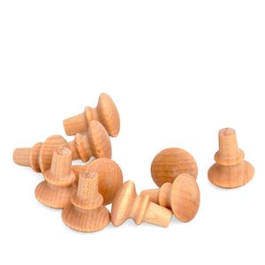 May include: A collection of unfinished wooden knobs and pegs, ideal for craft projects. The knobs have a rounded top and a cylindrical base, while the pegs are cylindrical with a small top. The wood has a natural, light brown color.