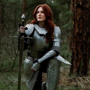May include: A woman in full plate armor kneels in a forest setting, holding a sword. She is wearing a silver helmet, breastplate, and leg armor. Her red hair is flowing down her back.
