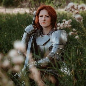 May include: A woman in silver armor sits in a field of tall grass, holding a sword. She has red hair and is looking directly at the camera.