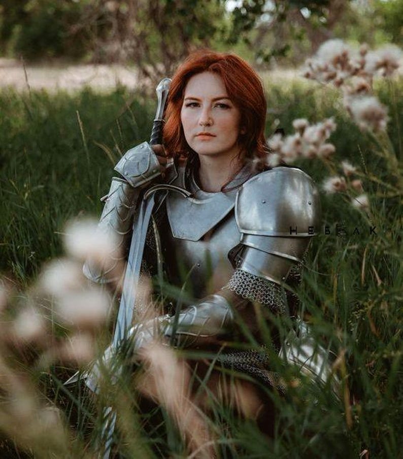 May include: A woman in silver armor sits in a field of tall grass, holding a sword. She has red hair and is looking directly at the camera.