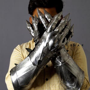 K&ouml;nnte beinhalten: Ein Paar silberne Metallhandschuhe mit komplizierten Designs und scharfen Spitzen an den Fingern. Die Handschuhe werden von einer Person getragen und bedecken ihre H&auml;nde und Unterarme.