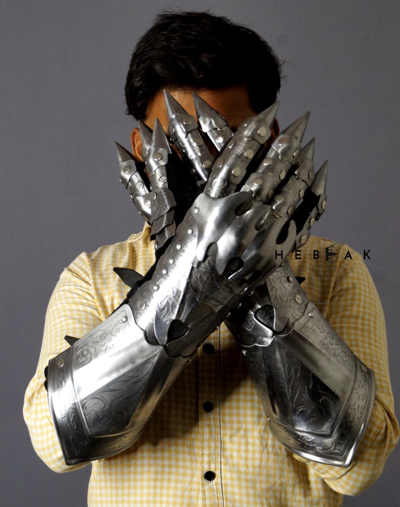 K&ouml;nnte beinhalten: Ein Paar silberne Metallhandschuhe mit komplizierten Designs und scharfen Spitzen an den Fingern. Die Handschuhe werden von einer Person getragen und bedecken ihre H&auml;nde und Unterarme.
