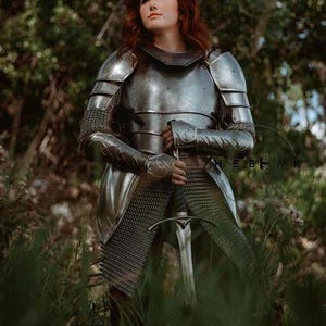 May include: A woman in full plate armor stands in a forest, holding a sword. She is wearing a chainmail skirt and a helmet with a visor. The armor is silver and has intricate details. The sword is long and sharp, with a silver blade and a black hilt.