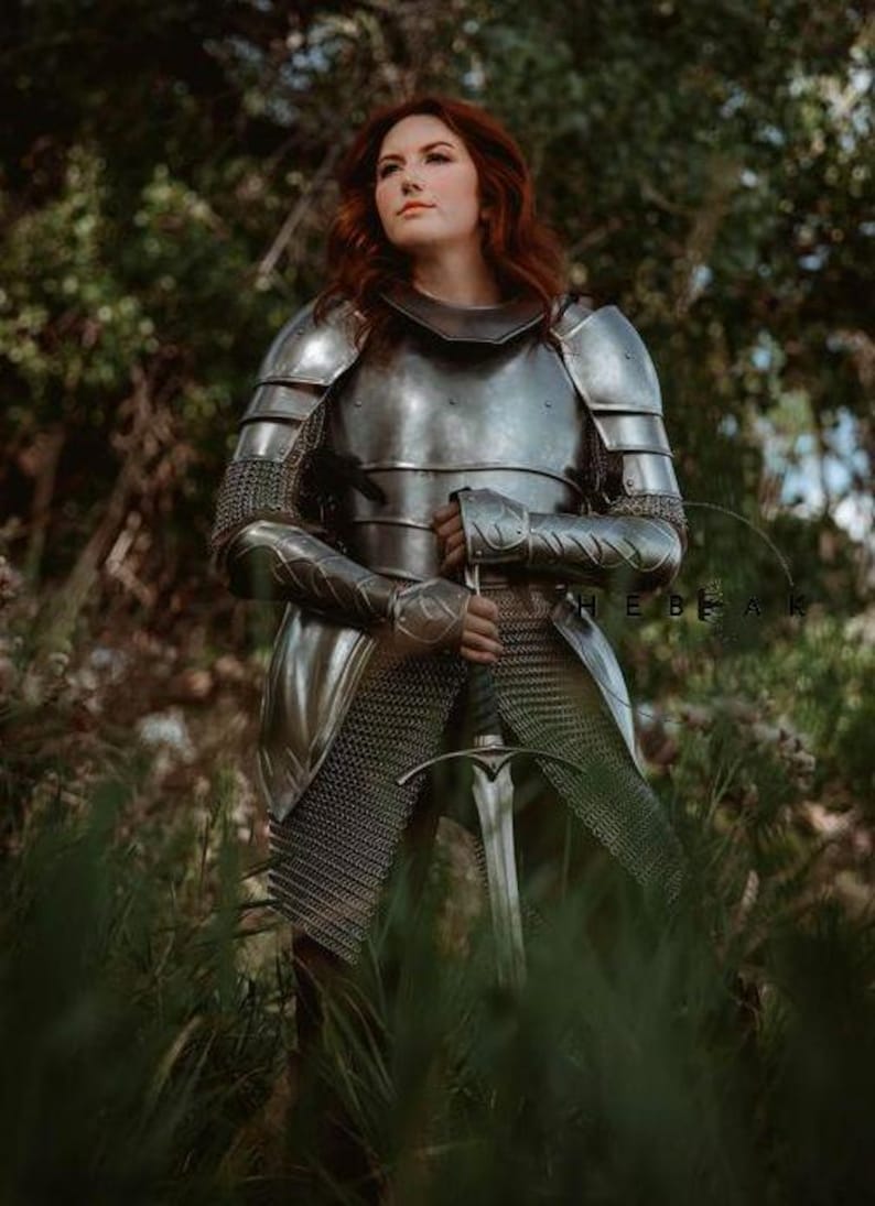 May include: A woman in full plate armor stands in a forest, holding a sword. She is wearing a chainmail skirt and a helmet with a visor. The armor is silver and has intricate details. The sword is long and sharp, with a silver blade and a black hilt.