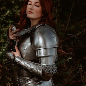 May include: A woman wearing a silver metal suit of armor with a sword in her hand. She has long red hair and is standing in a forest.