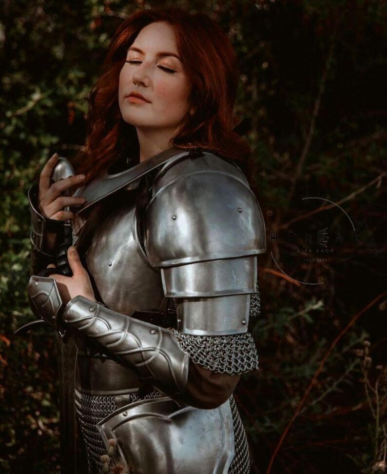 May include: A woman wearing a silver metal suit of armor with a sword in her hand. She has long red hair and is standing in a forest.