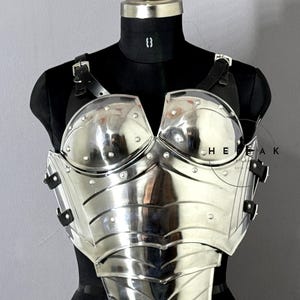 Puede incluir: Un peto plateado pulido con detalles remachados y correas de cuero, exhibido en un maniqu&iacute; negro. La armadura presenta un dise&ntilde;o curvo y es reflectante, con el texto "HEAK" visible.