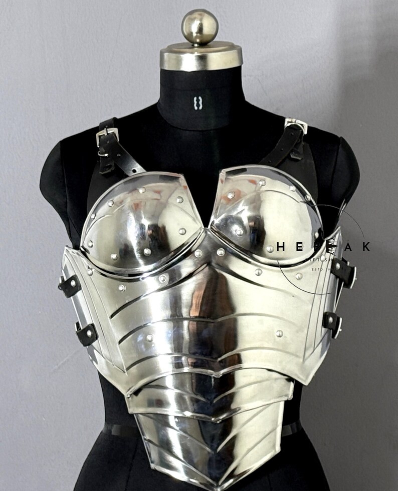 Puede incluir: Un peto plateado pulido con detalles remachados y correas de cuero, exhibido en un maniqu&iacute; negro. La armadura presenta un dise&ntilde;o curvo y es reflectante, con el texto "HEAK" visible.