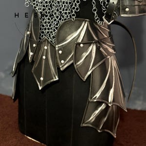 May include: A close-up of a black and silver fantasy armor set. The armor features a chainmail top, a leather belt with decorative plates, and a skirt-like lower section. The plates have a silver trim and are riveted. The armor is displayed on a black form.