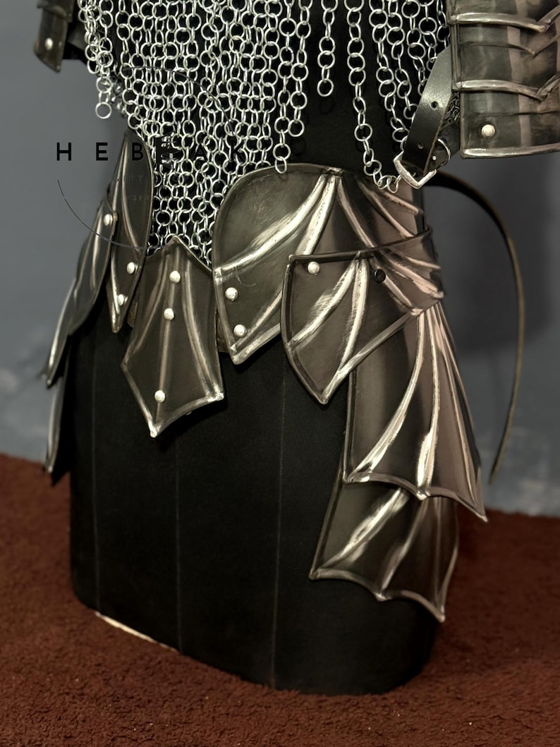 May include: A close-up of a black and silver fantasy armor set. The armor features a chainmail top, a leather belt with decorative plates, and a skirt-like lower section. The plates have a silver trim and are riveted. The armor is displayed on a black form.