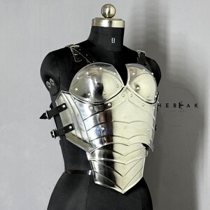 Puede incluir: Un peto plateado pulido con detalles de remaches y correas de cuero, exhibido en un maniqu&iacute; negro. La armadura presenta un dise&ntilde;o contorneado con placas en capas y una superficie reflectante, que evoca un tema medieval o de fantas&iacute;a.