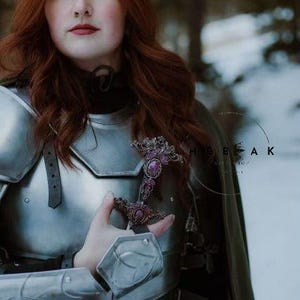 May include: A woman in silver armor with a green cloak, holding a sword with purple gemstones. The sword is adorned with intricate details.
