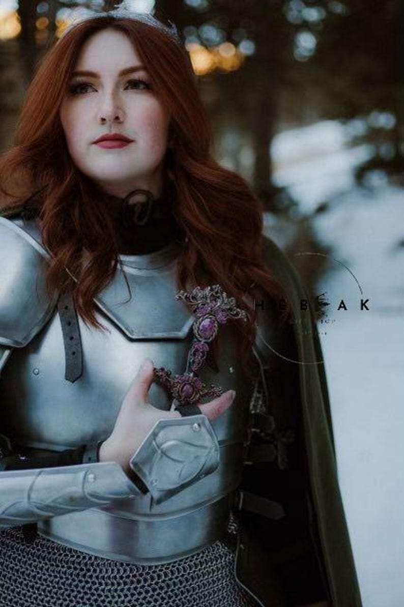 May include: A woman in silver armor with a green cloak, holding a sword with purple gemstones. The sword is adorned with intricate details.