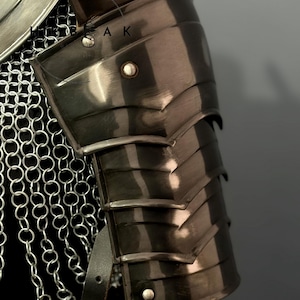 May include: Close-up of a section of medieval armor. The image shows a polished, dark metal pauldron with layered plates, riveted details, and a leather strap. Chainmail is visible in the background.