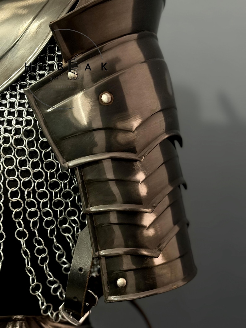 May include: Close-up of a section of medieval armor. The image shows a polished, dark metal pauldron with layered plates, riveted details, and a leather strap. Chainmail is visible in the background.