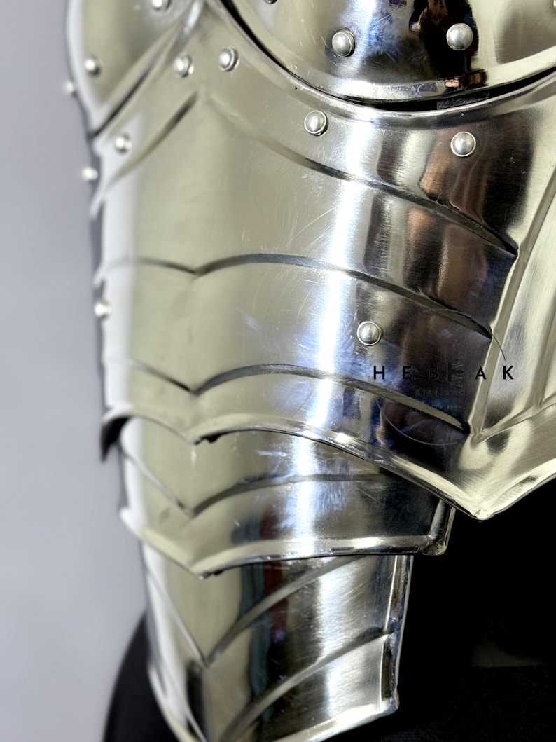 Puede incluir: Primer plano de una peto de metal plateado pulido. La armadura presenta placas curvas y en capas, y remaches. La superficie refleja la luz, destacando la artesan&iacute;a y el dise&ntilde;o de la armadura de estilo medieval.