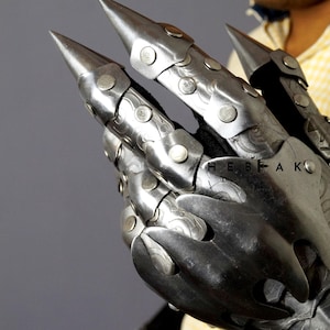 K&ouml;nnte beinhalten: Ein silberner Metall-Klauenhandschuh mit spitzen Fingern und Nieten. Der Handschuh wird von einer Person mit einem gelben Hemd getragen. Der Text "HEBEAK" ist auf dem Handschuh sichtbar.