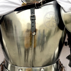 May include: A silver knight's armor with a leather strap and buckle across the chest. The armor has a cross embossed on the chest plate.