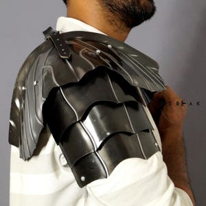 以下が含まれることがあります： 重ねられた鱗状のデザインが施された黒い金属製の肩当て。肩当ては左肩に装着され、革のストラップが付いている。