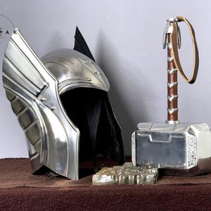 May include: A silver helmet with a winged design and a silver hammer with a brown leather strap. The helmet has a black fabric lining. The hammer has a silver head with intricate details.