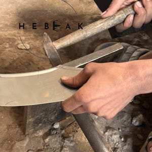 May include: A craftsman uses a hammer and anvil to shape a metal blade. The image shows the process of metalworking, with the text "HEBEAK GIFT SHOP ESTD 1994" in the upper left corner.