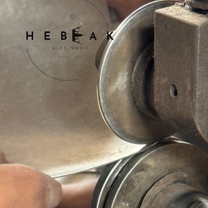 May include: Close-up of metalworking machinery shaping a sheet of metal. The image features the text "HEBEAK GIFT SHOP ESTD. 2014" in a circular logo. The machinery is in operation, with a hand guiding the metal.