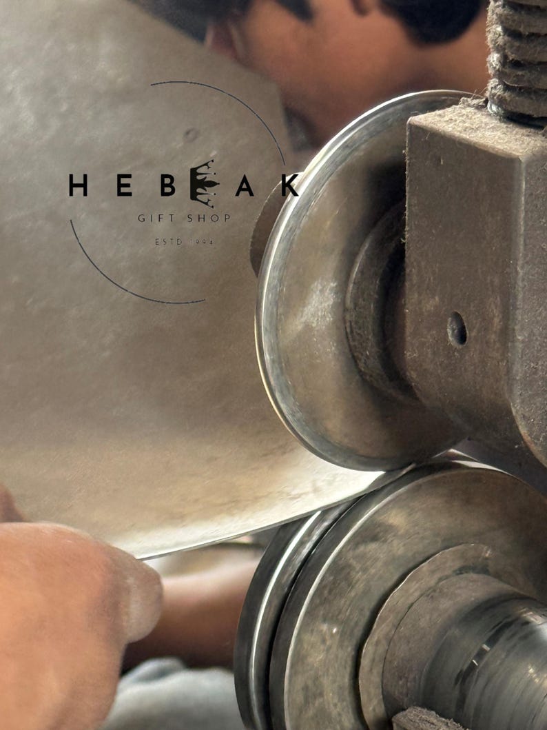 May include: Close-up of metalworking machinery shaping a sheet of metal. The image features the text "HEBEAK GIFT SHOP ESTD. 2014" in a circular logo. The machinery is in operation, with a hand guiding the metal.