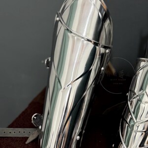May include: A pair of silver metal arm guards with a decorative design. The guards are made of metal plates that are joined together with rivets. The guards are designed to protect the wearer's arms from injury.