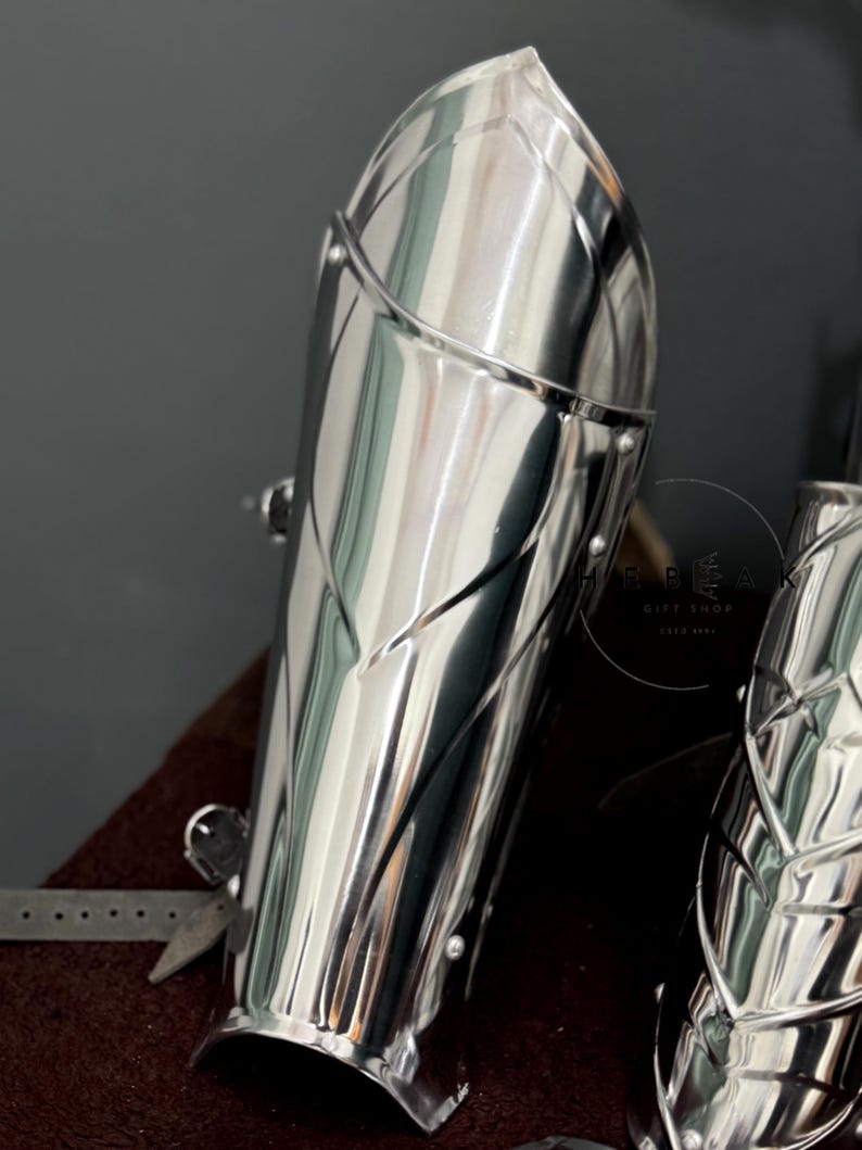 May include: A pair of silver metal arm guards with a decorative design. The guards are made of metal plates that are joined together with rivets. The guards are designed to protect the wearer's arms from injury.