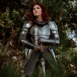 May include: A woman in full plate armor stands in a forest, holding a sword. She is wearing a chainmail skirt and a helmet with a visor. The armor is silver and has intricate details. The sword is long and sharp, with a black handle.