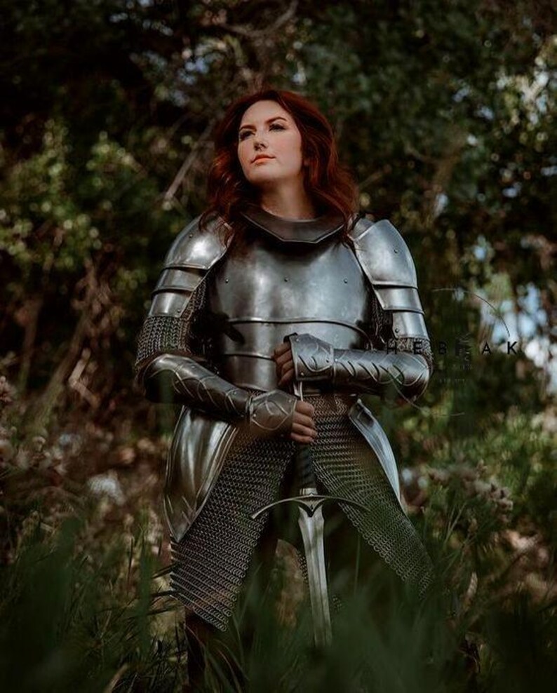 May include: A woman in full plate armor stands in a forest, holding a sword. She is wearing a chainmail skirt and a helmet with a visor. The armor is silver and has intricate details. The sword is long and sharp, with a black handle.