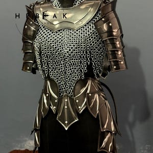 May include: A medieval-style armor set, displayed on a black mannequin. The armor features a silver breastplate, chainmail, and articulated shoulder and arm guards. The lower portion has layered, stylized metal plates. The armor is dark with silver accents.