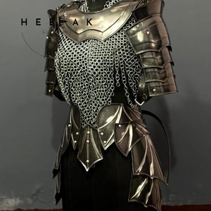 May include: A suit of dark metal armor displayed on a mannequin. The armor features layered plates, chainmail, and decorative elements. The text "HEE AK" is visible on the left side of the image. The armor is designed to protect the torso and arms.