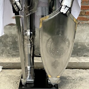 May include: A full suit of silver armor with a sword and shield. The shield has a gold trim and a design of a knight on horseback with a cross.