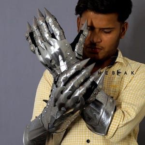 K&ouml;nnte beinhalten: Ein Paar silberne Metallhandschuhe mit spitzen Fingerkuppen und dekorativen Gravuren. Die Handschuhe werden von einer Person mit einem gelb-wei&szlig; karierten Hemd getragen.
