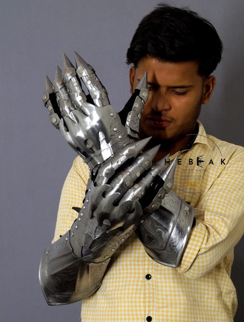 K&ouml;nnte beinhalten: Ein Paar silberne Metallhandschuhe mit spitzen Fingerkuppen und dekorativen Gravuren. Die Handschuhe werden von einer Person mit einem gelb-wei&szlig; karierten Hemd getragen.
