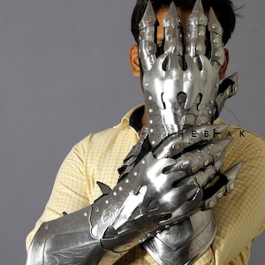 K&ouml;nnte beinhalten: Ein Paar silberne Metallhandschuhe mit krallenartigen Fingern und dekorativen Gravuren. Die Handschuhe werden von einer Person getragen und bedecken ihre H&auml;nde und Unterarme.