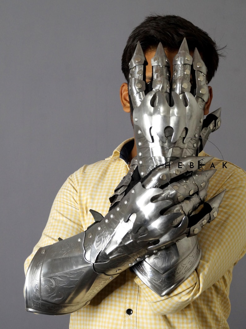 K&ouml;nnte beinhalten: Ein Paar silberne Metallhandschuhe mit krallenartigen Fingern und dekorativen Gravuren. Die Handschuhe werden von einer Person getragen und bedecken ihre H&auml;nde und Unterarme.
