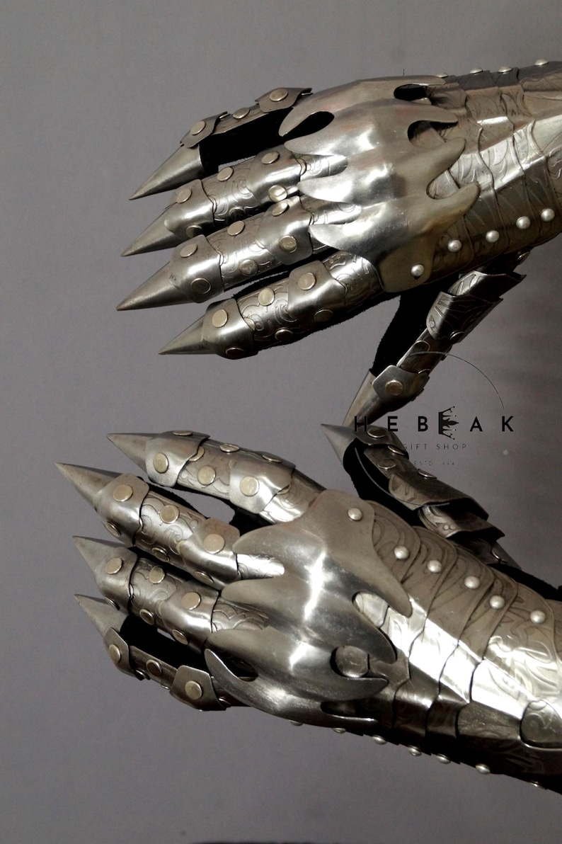 K&ouml;nnte beinhalten: Ein Paar silberne Metallhandschuhe mit spitzen Krallen und dekorativen Details. Die Handschuhe bestehen aus Metallplatten und haben eine strukturierte Oberfl&auml;che. Die Handschuhe werden an beiden H&auml;nden getragen und werden in einer Greifposition zusammengehalten.