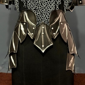 May include: Close-up of a black and silver medieval armor set. The armor features layered leather and metal plates, with chainmail detailing at the top. The design includes decorative elements and rivets, showcasing a detailed craftsmanship.