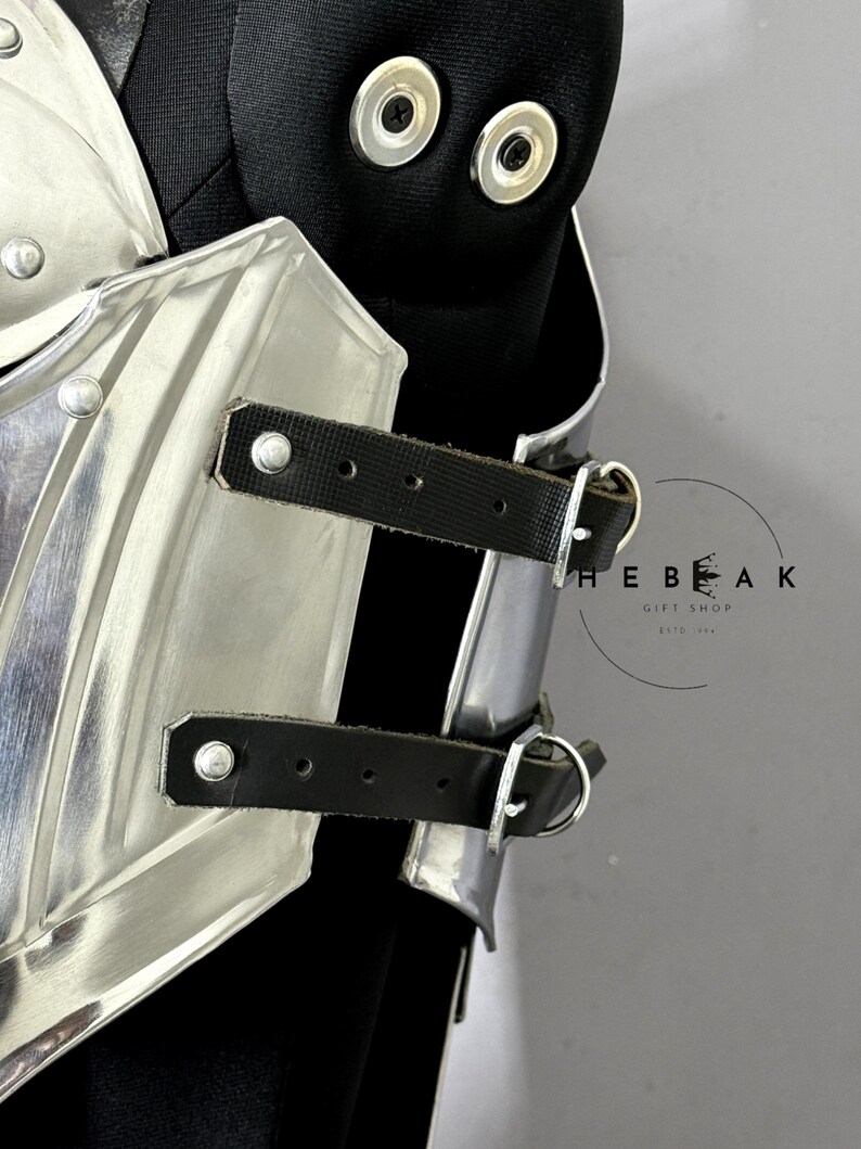 Puede incluir: Primer plano de una pieza de armadura de hombro de metal plateado con correas de cuero negro y remaches. La armadura est&aacute; sujeta a una prenda negra. La imagen incluye el texto "THE BEAK GIFT SHOP ESTD 2016".