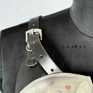 Puede incluir: Primer plano de una correa de hombro de cuero negro con hebilla y remaches plateados. La correa est&aacute; sujeta a una pieza de armadura con un acabado met&aacute;lico pulido. La armadura parece ser parte de un disfraz medieval o de fantas&iacute;a.