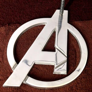 May include: A silver metal pendant shaped like the Avengers logo, featuring a stylised "A" inside a circle. The pendant is hanging from a black cord.