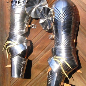 May include: Highly polished steel armour components, including leg and arm guards, and small circular shields. The armour is displayed against a wooden background. The metal reflects light, highlighting the craftsmanship and historical design.