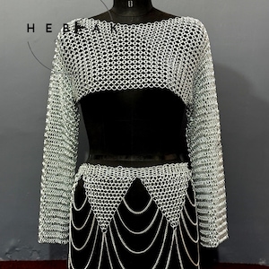 May include: A silver chainmail top and skirt set displayed on a black mannequin. The top is a cropped, long-sleeved design. The skirt features a layered chain design. The set is made of small, interlocking metal rings.