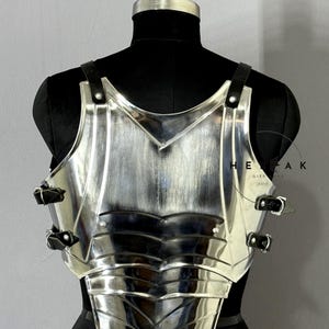 Puede incluir: Una armadura de pecho plateada pulida con correas y hebillas de cuero negro. La armadura se exhibe en un maniqu&iacute; negro. La armadura tiene un dise&ntilde;o en capas con un escote en forma de V y est&aacute; dise&ntilde;ada para proteger el pecho.