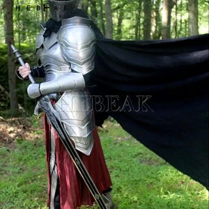 May include: A full body suit of silver armor with a black cape and a sword. The armor has a helmet with a visor and a skirt. The person is standing in a forest.