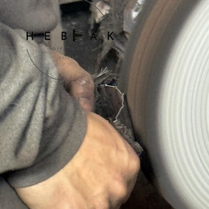 May include: Close-up of a craftsman working on a metal object with a grinding wheel. The image shows the hands of the artisan holding the metal piece against the spinning wheel. The text "HEBEAK GIFT SHOP ESTD" is visible.